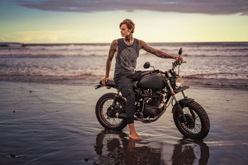 Obraz na płótnie Canvas tattooed biker sitting on motorcycle on ocean beach and looking away