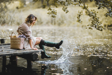 Woman in rubber boots sitting on wooden bridge near lake