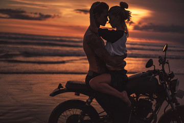 Obraz na płótnie Canvas shirtless boyfriend hugging girlfriend on motorcycle at beach during sunset