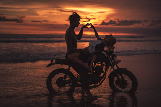 passionate couple making heart with fingers on motorcycle at beach during sunset