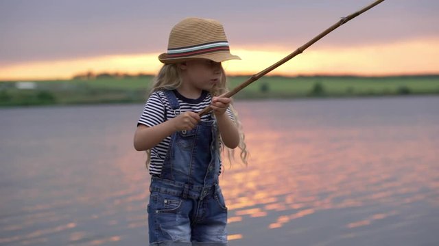 Portrait slow motion of little girl 7-8 with blonde long hair wearing straw hat holding wooden rod and fishing on lake or river during sunset in countryside