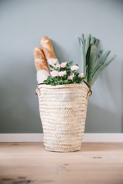 A rush or wicker basket with light pink roses, with two baguettes and a couple of leeks.
