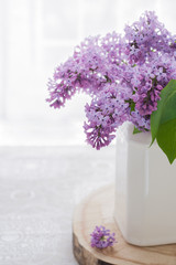White ceramic pot with purple lilac is standing on wooden round board, on linen tablecloth. High key.