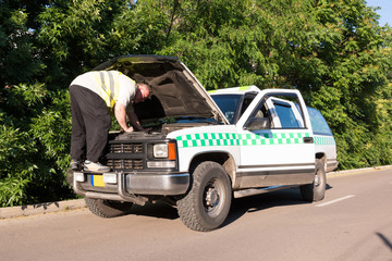 A mature man repairing his pickup truck with opened hood in a sunny outdoor environment.