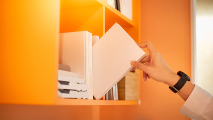 male hand choosing and picking white book on orange bookshelf in public library, education research...