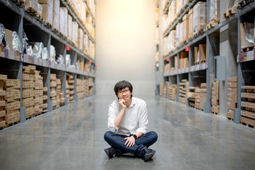 Young Asian man sitting between cardboard bon shelves in warehouse choosing what to buy, shopping warehousing concept