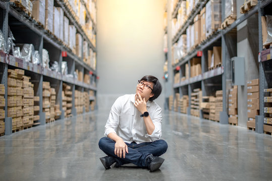 Young Asian man sitting between cardboard bon shelves in warehouse choosing what to buy, shopping warehousing concept