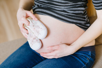 A caucasian pregnant woman sitting on sofa and holding a little shoes