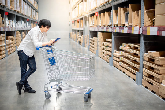 Young Asian man standing with cart checking the shopping list and looking for product in warehouse wholesale, shopping warehousing concept