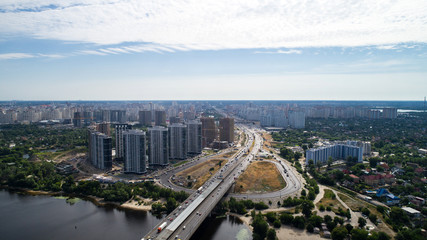 South Bridge. Aerial view of many buildings. The Dnieper River. Summer. The sun. Day. Green trees. High-rise buildings. Road. Cars. Infrastructure. Sky. The clouds. Kyiv.