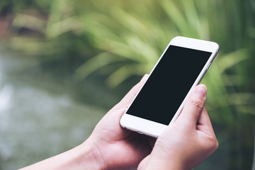 Mockup image of a woman holding white smart phone with blank black desktop screen with blur green nature background