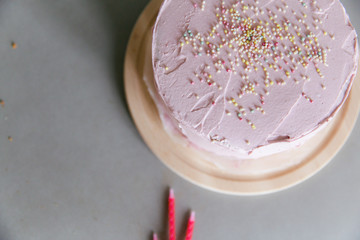 A homemade pink birthday cake with sprinkles layer out on a round wooden cutting board on a grey ceramic kitchen counter top with three birthday candles.