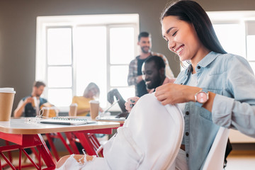 happy young student girl opening backpack at classroom with blurred mates on background
