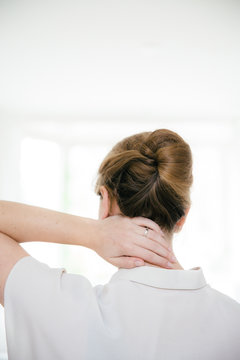 A woman with a fancy updo is touching her neck. She is seen from the back and is wearing a short sleeved beige blouse.