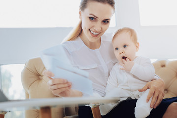 Have a look. Joyful blonde embracing her kid while looking at sheet of paper