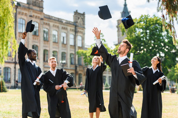 Young happy graduated students throwing up graduation caps in university garden