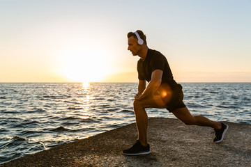 athletic adult man in headphones doing one legged squats during training on seashore