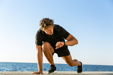 handsome adult sprint runner looking at fitness tracker while standing in start position for run on...