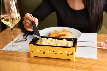 the girl tastes cheese raclette with a glass of white wine in a cafe. swiss food. cheese raclette with mushrooms on coupelles - special small skillet for a raclette
