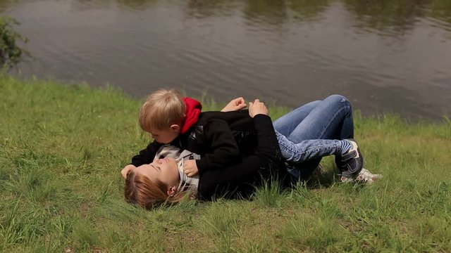 A happy mother with her son lies on the green grass on the river bank in the spring. Mom and son are walking in the park, they lie on the ground hugging and kissing.
