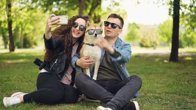 Pretty woman is taking selfie with her boyfriend and adorable dog using smartphone while resting in the park on the grass. Humans and animals are wearing sunglasses.