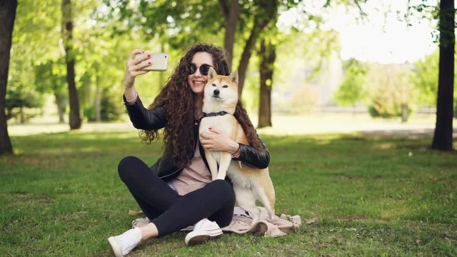 Cheerful blogger pet owner is taking selfie with her dog using smartphone, human and animal are sitting on lawn in the park and posing, woman is caressing and kissing dog.