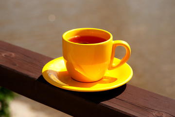 A bright cup of tea for relaxing. A yellow cup of tea against the background of the water in the river. Tea in the sun on the wooden railing of the cafe.