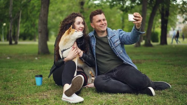 Handsome young man is taking selfie with his girlfriend who is holding pedigree dog, guy is kissing girl then watching photos on screen, people are laughing.