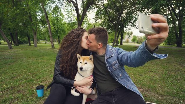 Cheerful guy is taking selfie holding smartphone and kissing his wife while she is holding adorable dog and kissing it. Relationship, love, romance and technology concept.