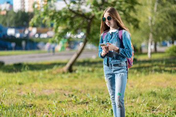 Girl schoolgirl. Summer in nature. In hands smartphone. She is wearing jeans clothes. Behind backpack. Free space for text. The concept communication on Internet. Writes messages social networks.