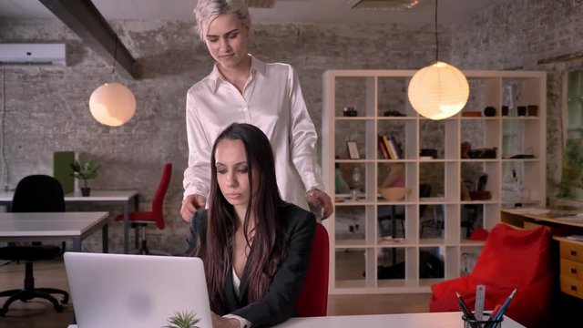 Young blonde lesbian massaging woman in modern office during working, woman flirting with woman, seductive and attractive