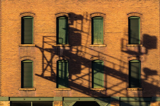Green window shutters on a red brick building with an interesting shadow and symetry.