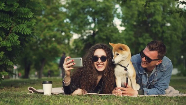 Young woman is taking selfie in the park lying on grass with her boyfriend and pet dog, adorable animal is sneezing and licking its nose, people are laughing.