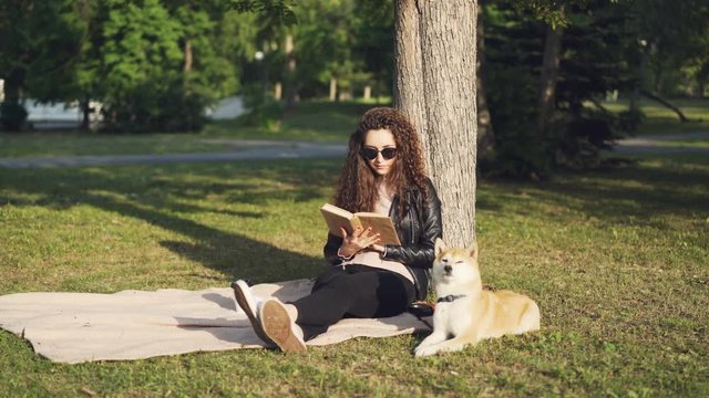 Attractive girl student is reading book sitting on plaid under tree in city park with her puppy lying near and enjoying sunlight. Hobby, leisure and animals concept.