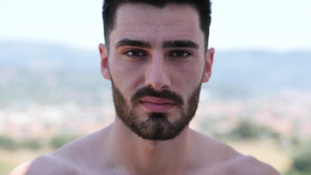 Handsome shirtless athletic young man outdoor standing on balcony in a hot summer day, looking at camera and smiling