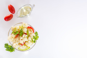 Fresh salad of cauliflower with vegetables and herbs in a glass bowl along with a glass nipple and tomatoes on a white background. Top view. Copy space