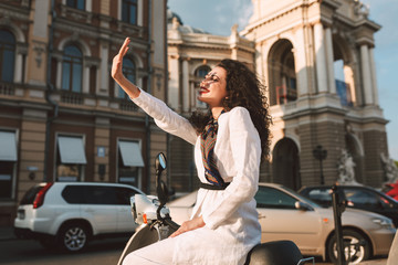 Naklejka premium Pretty smiling woman with dark curly hair in white costume sitting on moped and happily covering her face with hand from sunlights with city view on background