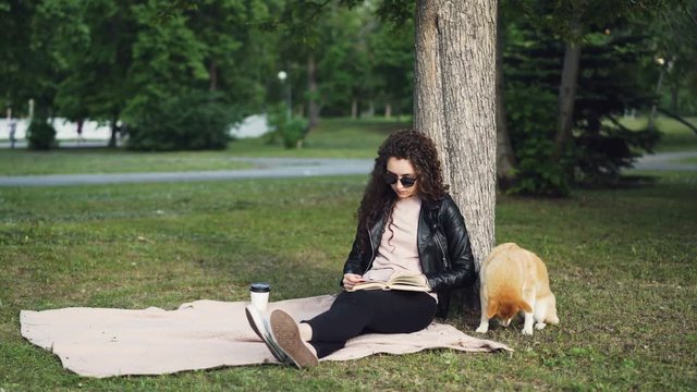 Relaxed girl is reading book and drinking takeaway coffee sitting on lawn in park while her cute dog is smelling ground and air sitting under tree. Leisure and pets concept.