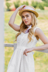 attractive blonde woman in wicker hat and white dress leaning at railing and looking at camera