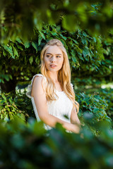 selective focus of beautiful blonde girl in white dress posing near green leaves