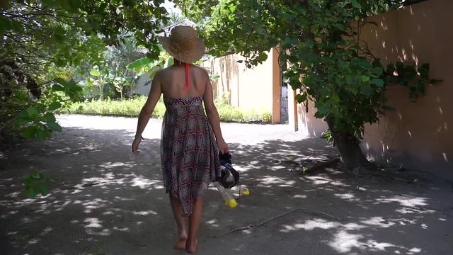 junges Mädchen in buntem Kleid und Strohhut spaziert auf Sandweg zwischen tropischen Pflanzen mit Schnorchelmasken 2