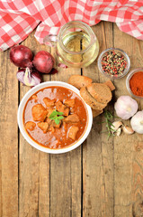 View from above of pork goulash with pieces of meat in a bowl, oregano, garlic, pepper, onion, jug with oil and red checkered tablecloth in the background - vertical photo
