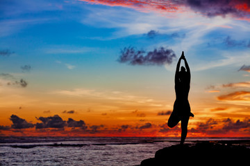 Sunset meditation silhouette. Active woman stand in yoga pose on beach rock to keep fit and health....