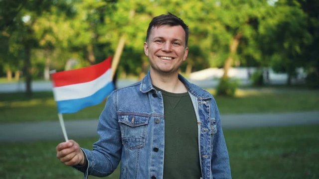 Slow motion portrait of joyful guy happy tourist waving flag of the Netherlands, smiling and looking at camera. National pride, visiting countries and people concept.