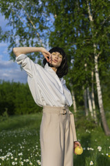 Beautiful stylish girl wearing a trendy white shirt, beige trousers and a black wig holds a green apple in sunlight on a background of a blue cloudy sky.