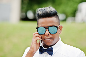 Close up portrait of stylish arabian man at sunglasses and bow tie posed outdoor and speaking on mobile phone. Arab model boy.