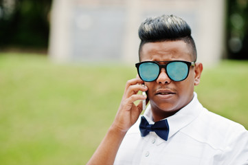 Close up portrait of stylish arabian man at sunglasses and bow tie posed outdoor and speaking on mobile phone. Arab model boy.