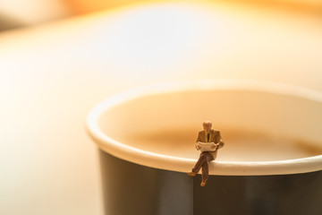 Business and reading concept. Businessman miniature figure sitting and reading a book on top of paper cup of hot coffee latte.