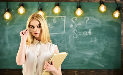 Woman with book starts lesson, gazes at audience while taking off eyeglasses. Teacher looks confident in eyeglasses, stand in classroom, chalkboard on background. Attractive lecturer concept.