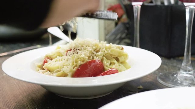 Women's hands grating parmesan cheese on a ready pasta with tomatoes, almonds and pesto sauce on a white plate. 4K